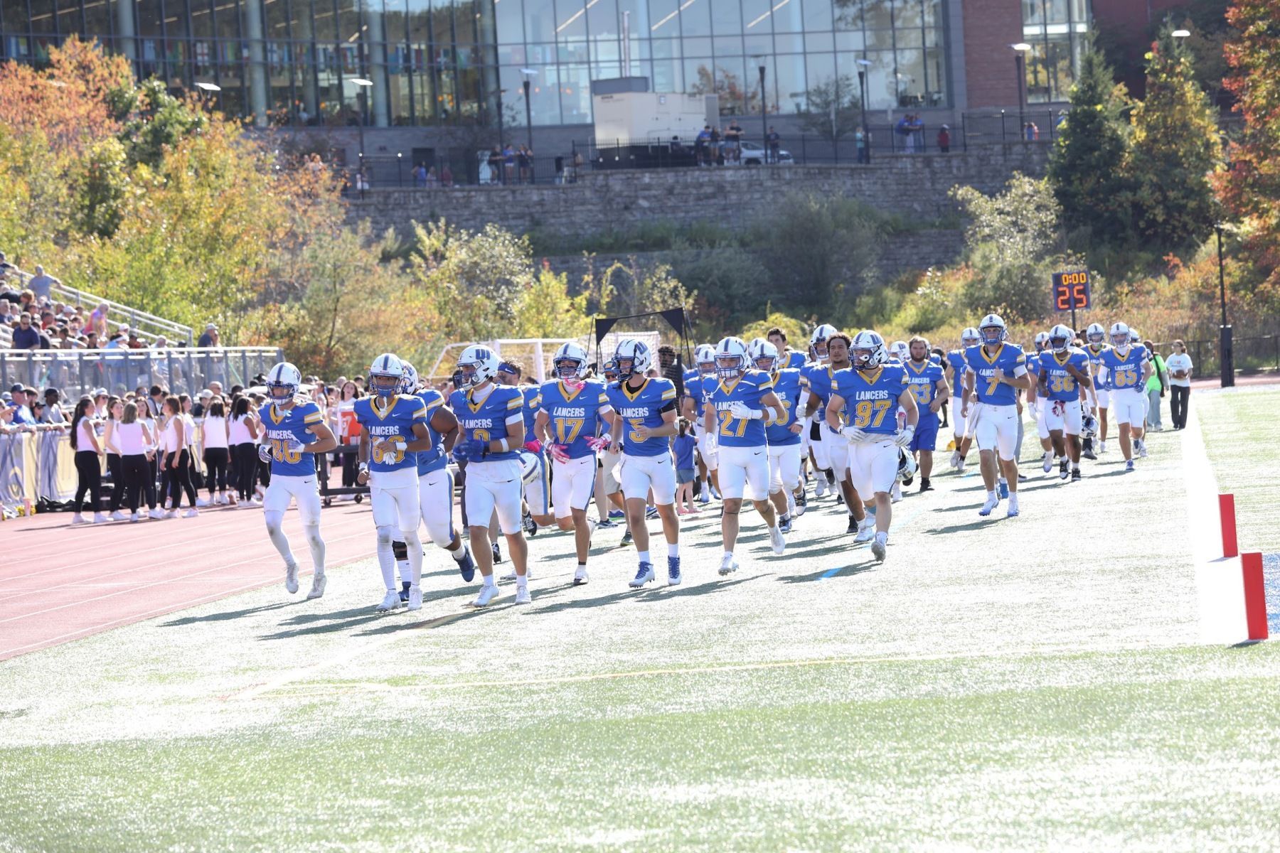 A college football team in blue and white uniforms runs onto the field, with spectators watching from the stands in the background.