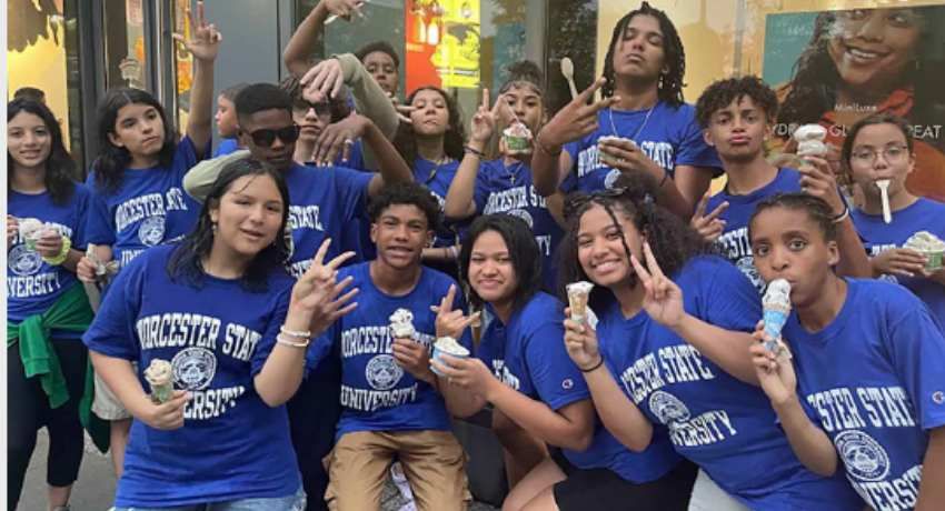 A group of teens wearing blue Worcester State University shirts pose together outdoors, holding ice cream cones and making hand signs.