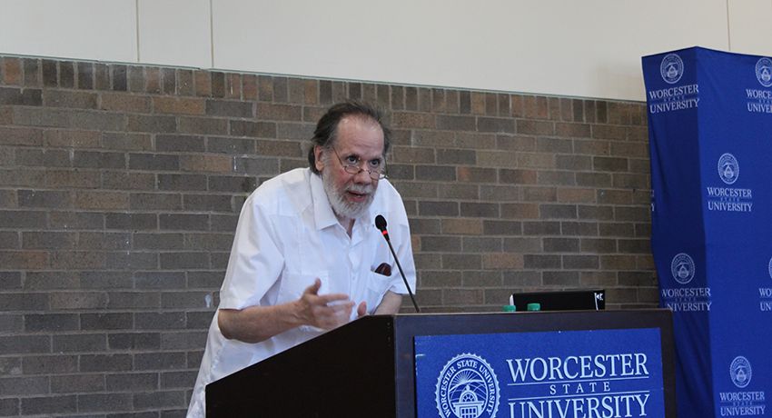 A man speaks at a podium with a Worcester State University sign, standing in front of a brick wall and university-branded backdrop.
