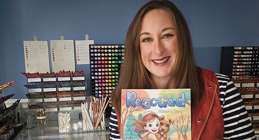 A woman smiles while holding a children's book titled "Rescued: Watson's Story" in an art studio with colored pencils and brushes in the background.