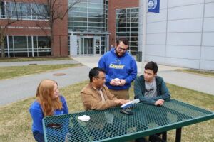 Four people sit and stand around a green outdoor table on a campus, engaged in conversation with papers and phones visible on the table.