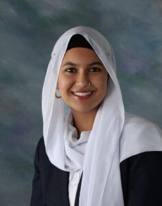 A woman wearing a white hijab, black blazer, and white shirt is smiling at the camera against a blurred studio background.