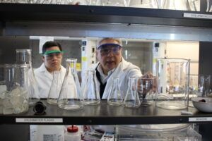 Two people in lab coats and safety goggles are viewed through a shelf of Erlenmeyer flasks and beakers in a laboratory setting.
