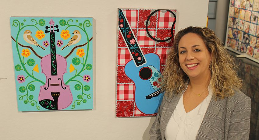 A woman stands beside two colorful paintings of guitars displayed on a gallery wall.