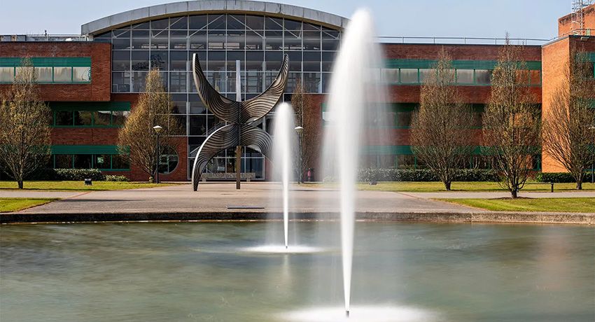A pond with two fountains in front of a modern brick and glass building, featuring a large abstract metal sculpture near the entrance.