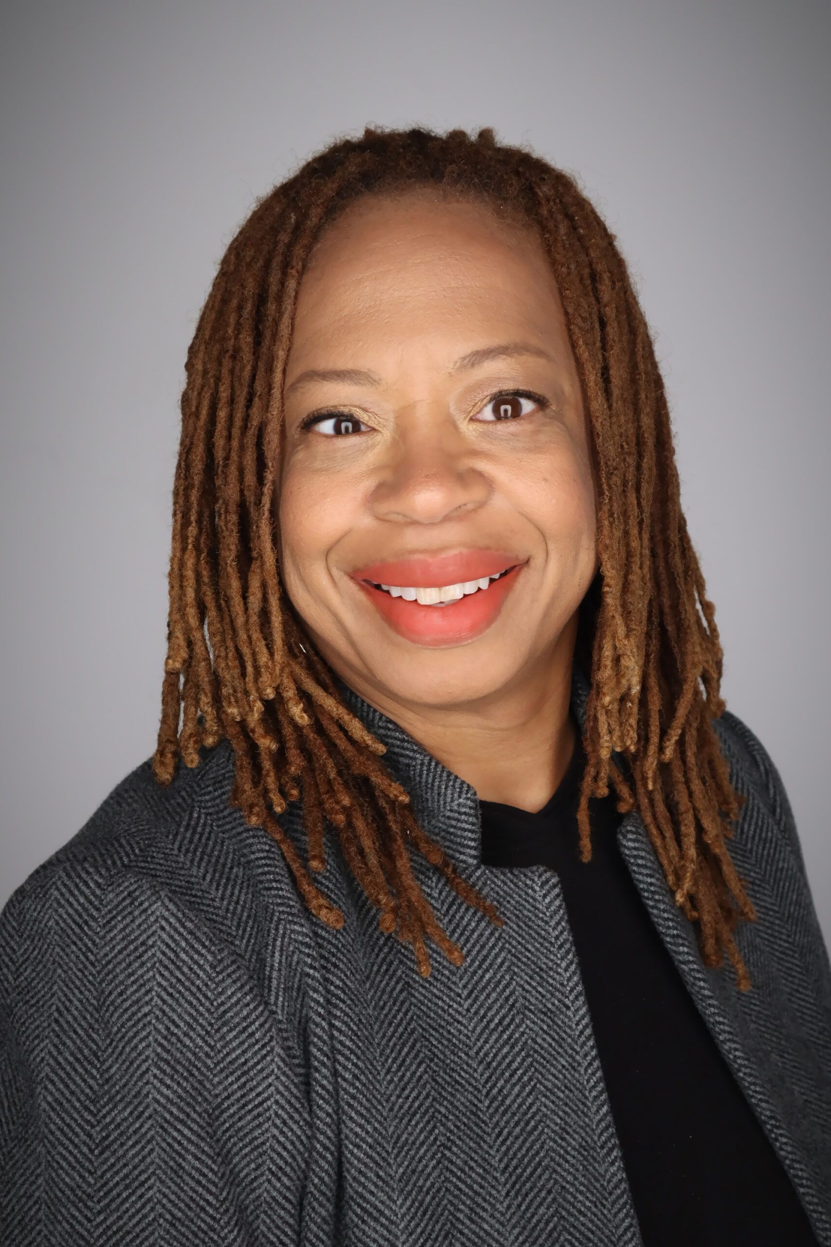 A person with shoulder-length dreadlocks, wearing a dark blazer and smiling, is positioned against a neutral gray background.