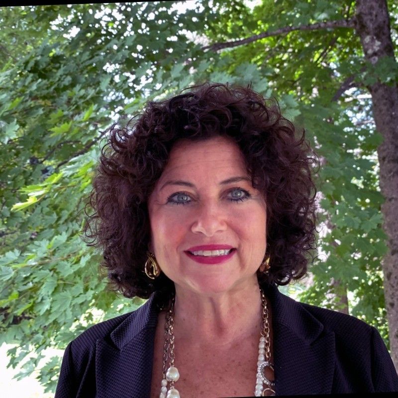 Woman with curly dark hair wearing a black blazer and necklace, smiling outdoors with green leafy trees in the background, embodies the spirit of University Advancement.