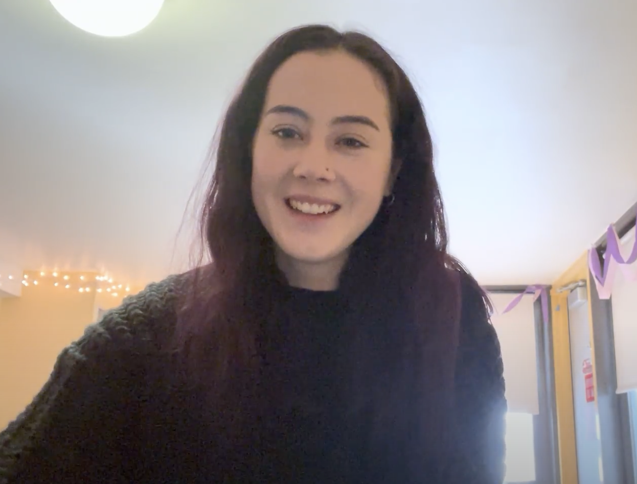 A person with long dark hair, who is pursuing a master in occupational therapy, is smiling in a brightly lit room adorned with string lights and purple decorations.