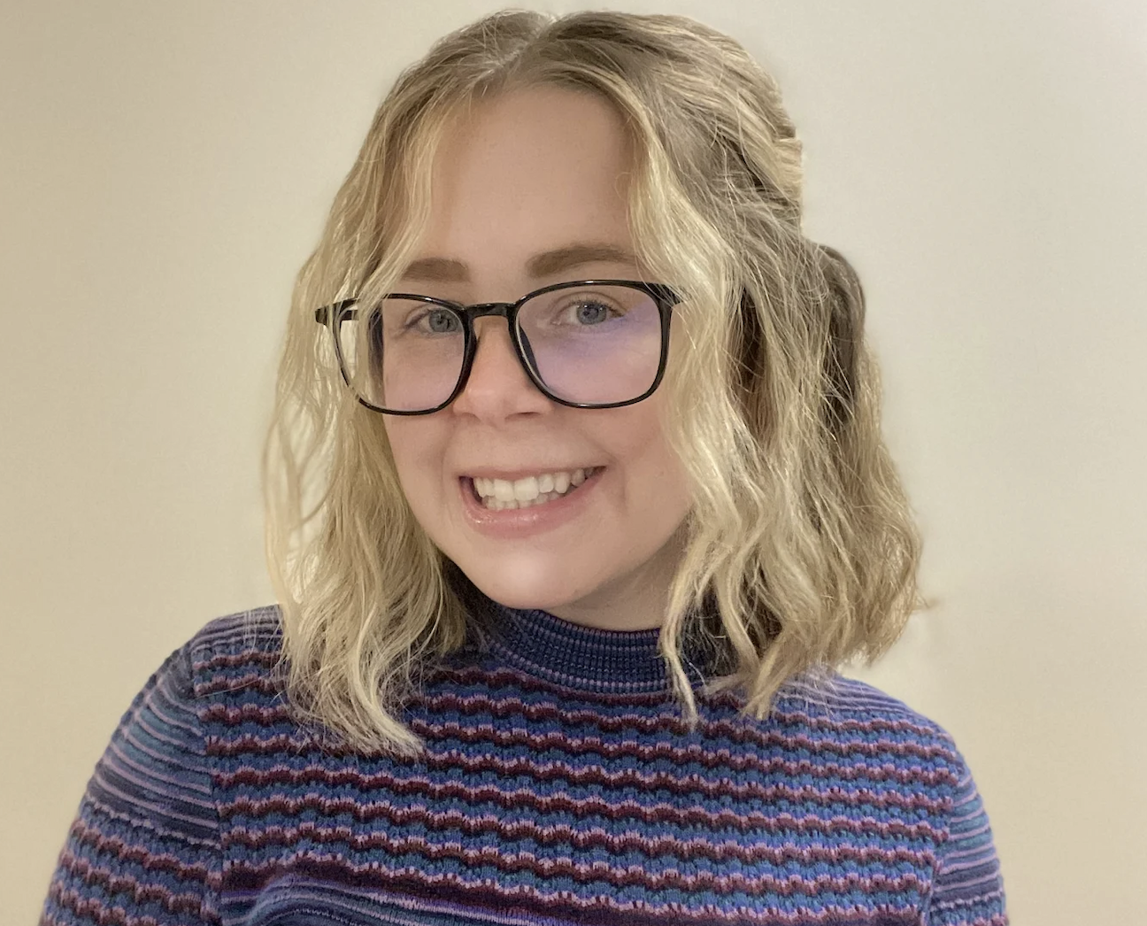 A smiling person with shoulder-length blonde hair and glasses wears a striped, textured, high-neck sweater against a neutral background. Their confident demeanor reflects their expertise as a master of occupational therapy.