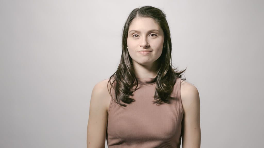 A woman with long dark hair in a sleeveless turtleneck top stands against a plain gray background, presenting the composed demeanor of someone who might master occupational therapy, facing forward with a neutral expression.