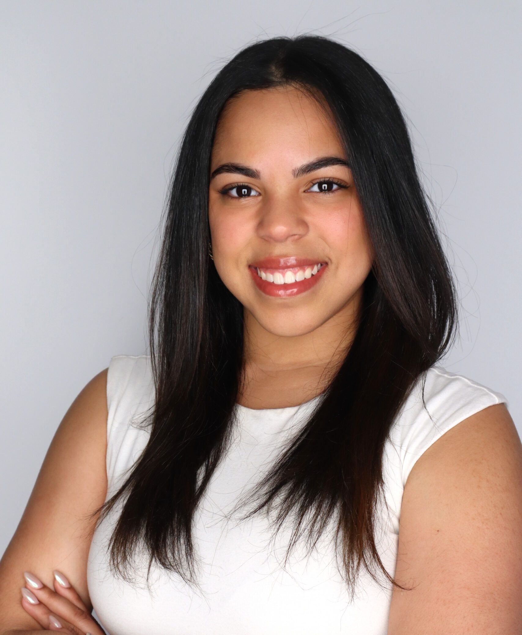 A young woman with long dark hair smiles with her arms crossed, wearing a sleeveless white top as a student of a master of occupational therapy.