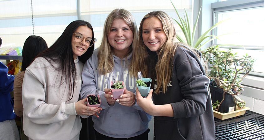 Three young women, one a biology major, smiling and holding small potted succulents inside a room with large windows and other plants in the background.
