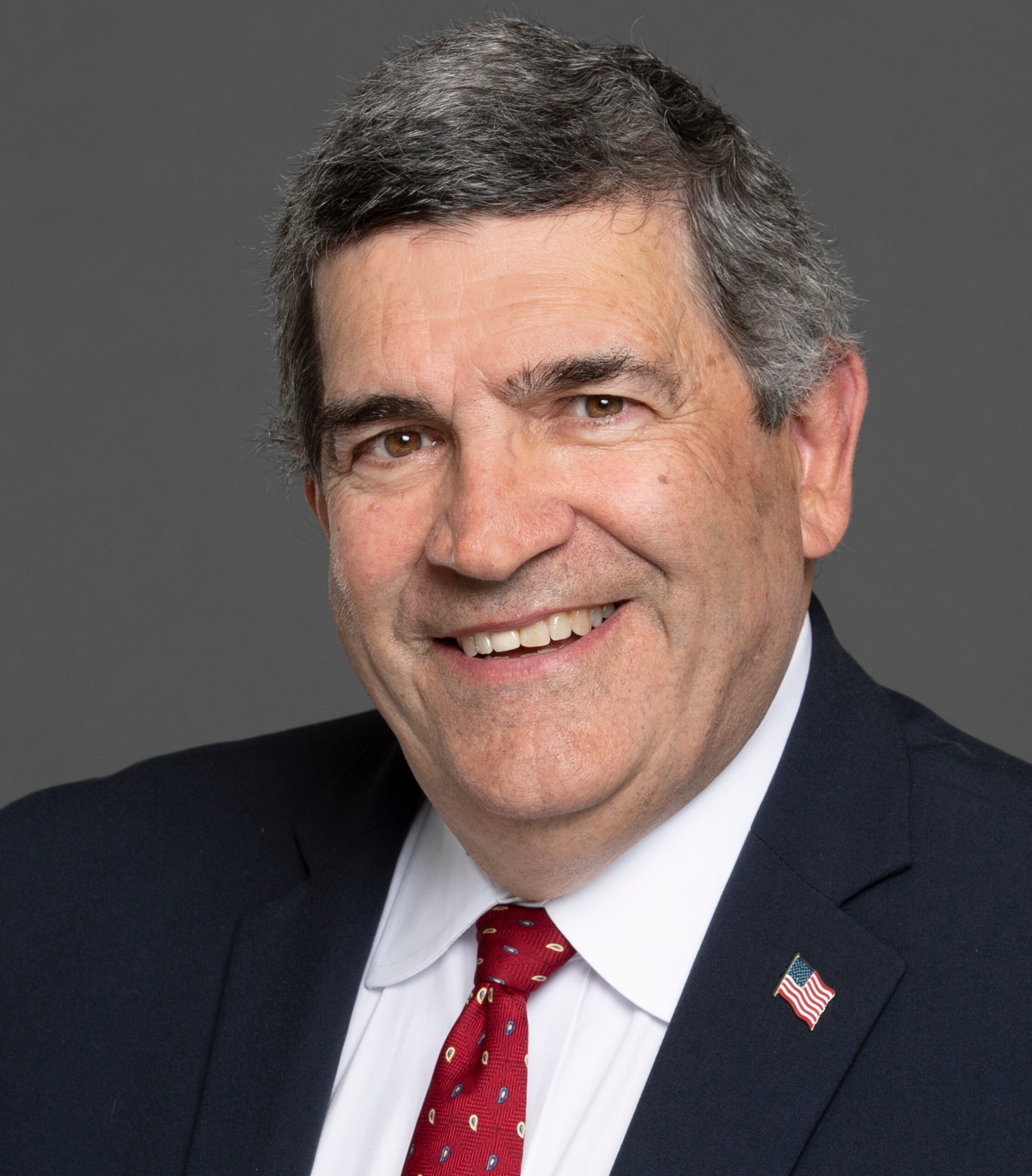 A man in a suit, embodying professionalism, smiles confidently against a gray background. His tie and lapel pin with an American flag design reflect his commitment to excellence in the field of criminal justice.