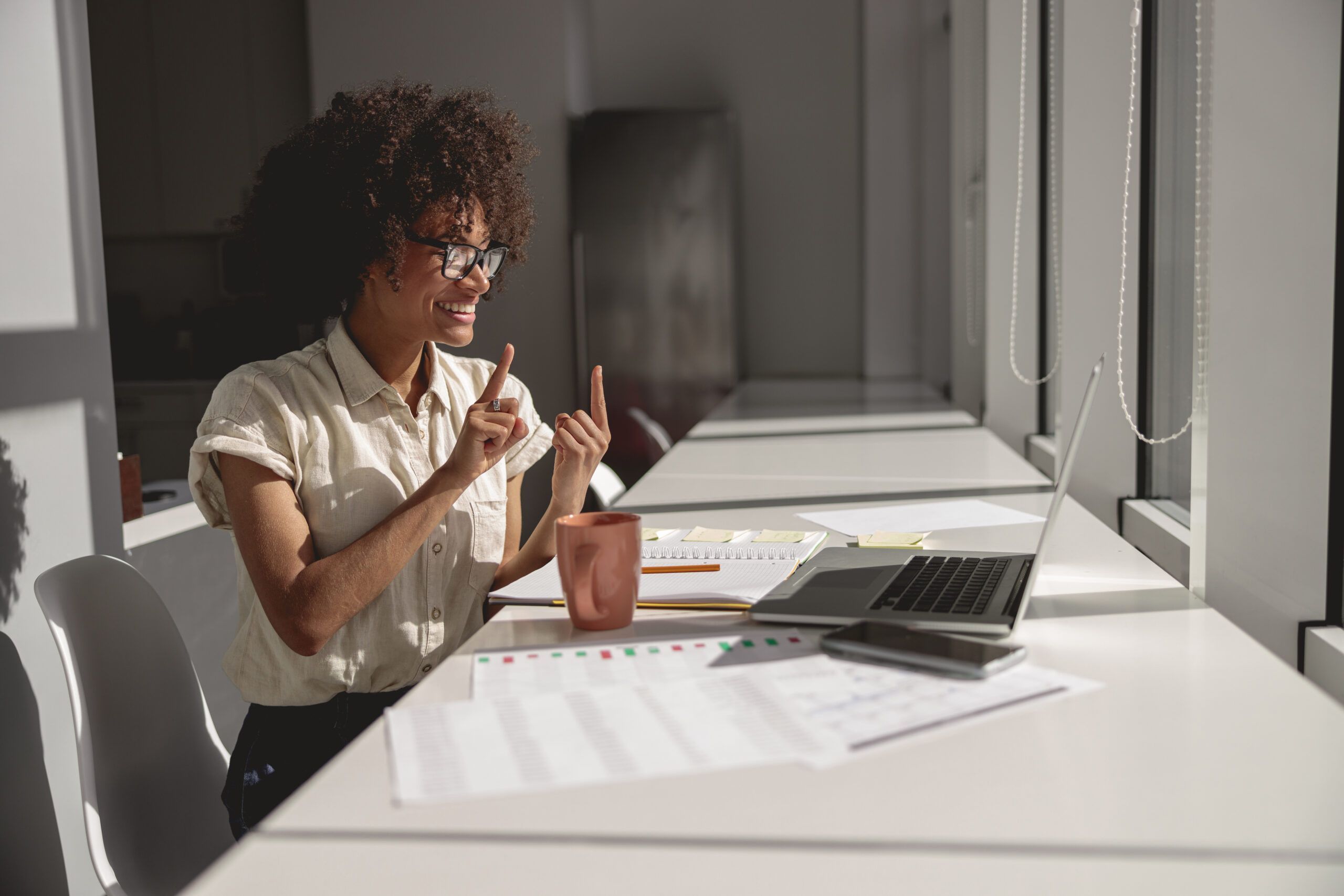 A Communication Sciences and Disorders major with curly hair and glasses signs in front of a laptop at a desk cluttered with papers and a mug, all illuminated by the bright lighting of the room.