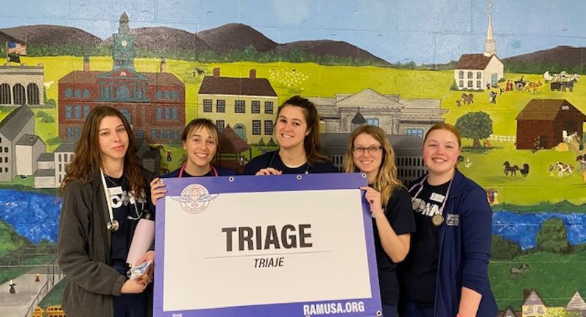 Five individuals, including a nursing major, stand in front of a painted mural, holding a large sign that reads "TRIAGE / TRIAJE" with the website "RAMUSA.ORG" at the bottom. All are wearing dark-colored shirts.