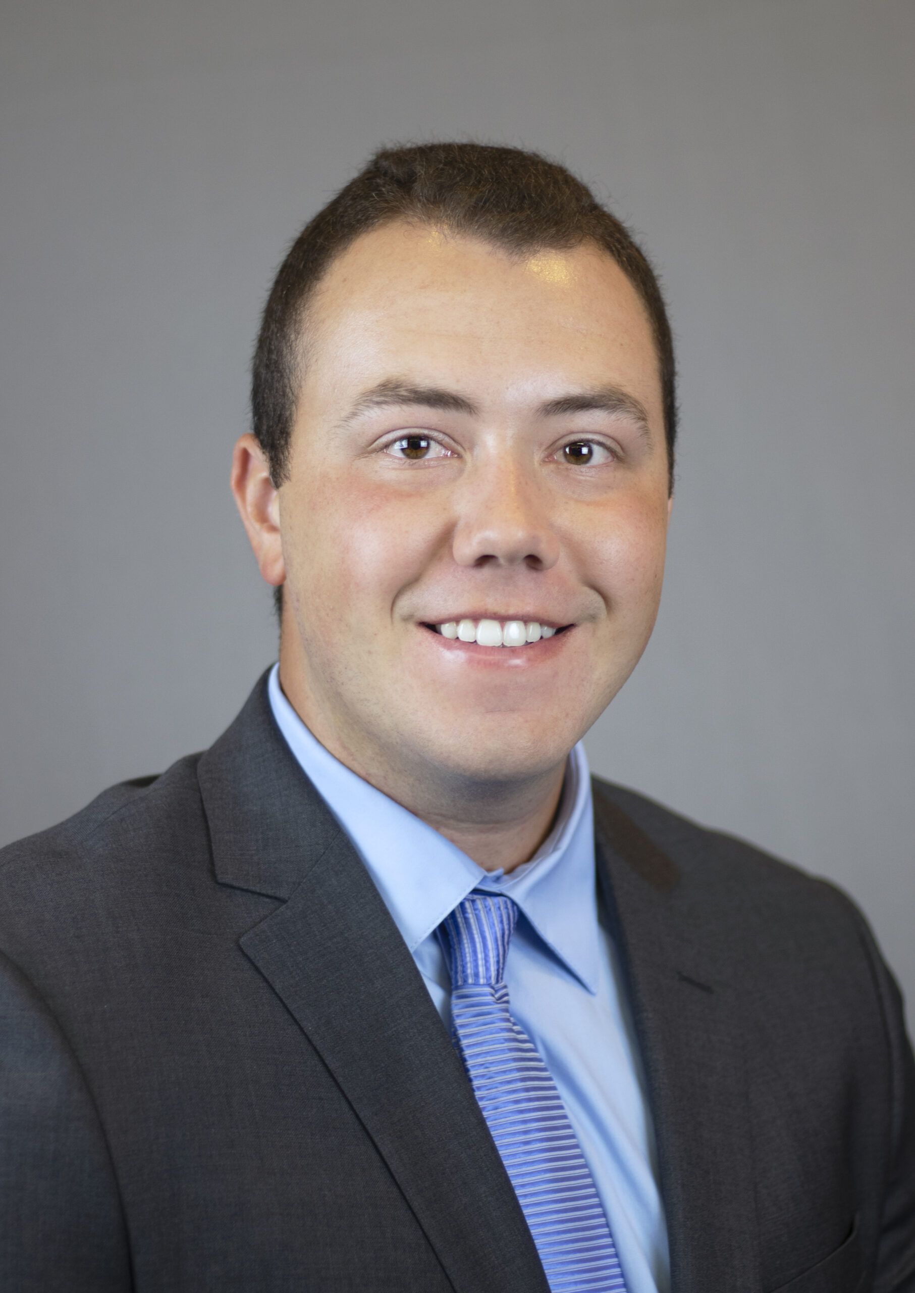 A person wearing a suit with a blue shirt and striped tie smiles confidently, embodying the integrity of the criminal justice system, in front of a gray background.