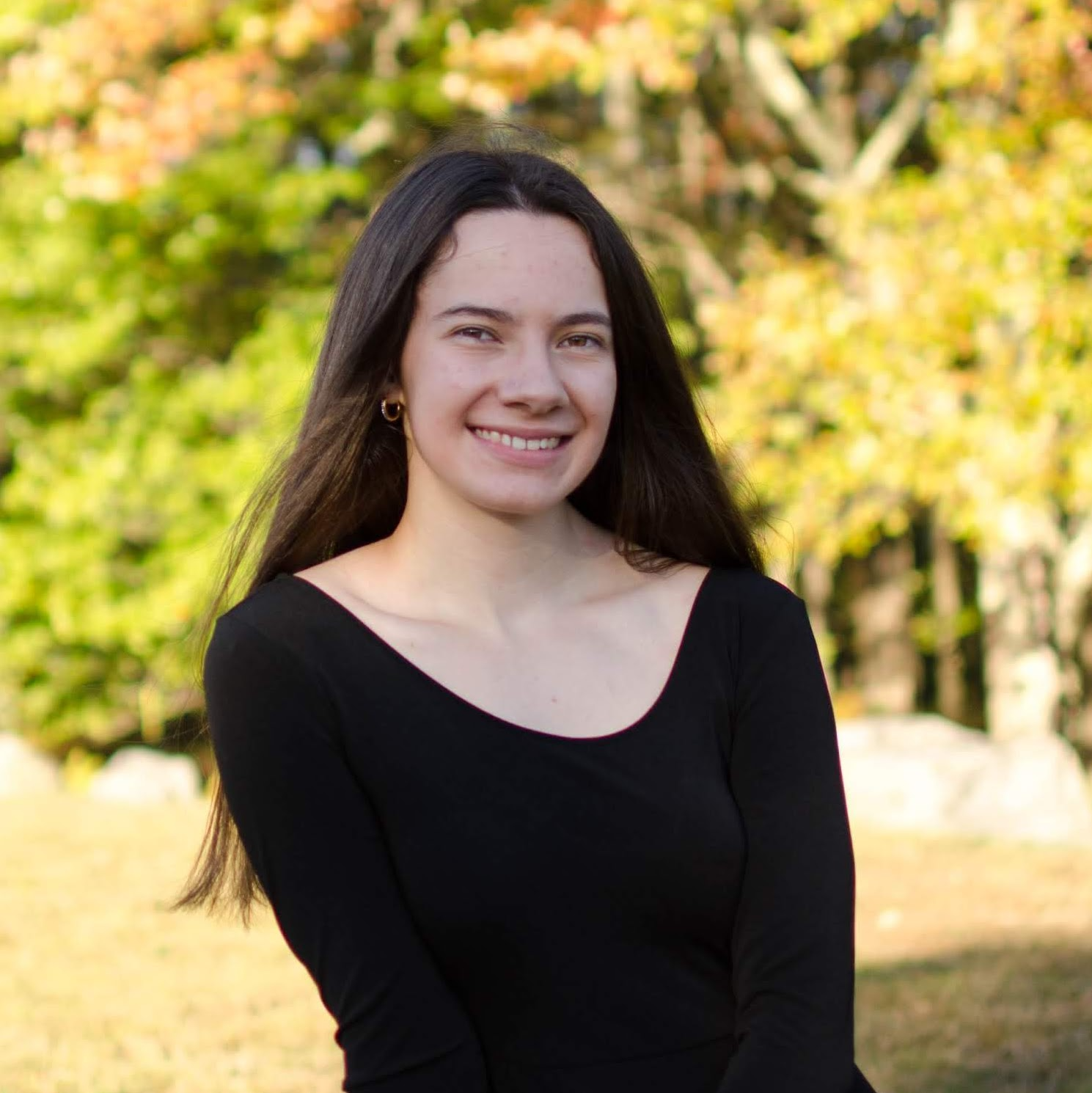 A young woman with long brown hair, wearing a black top, sits outdoors in front of trees with autumn foliage, smiling at the camera.