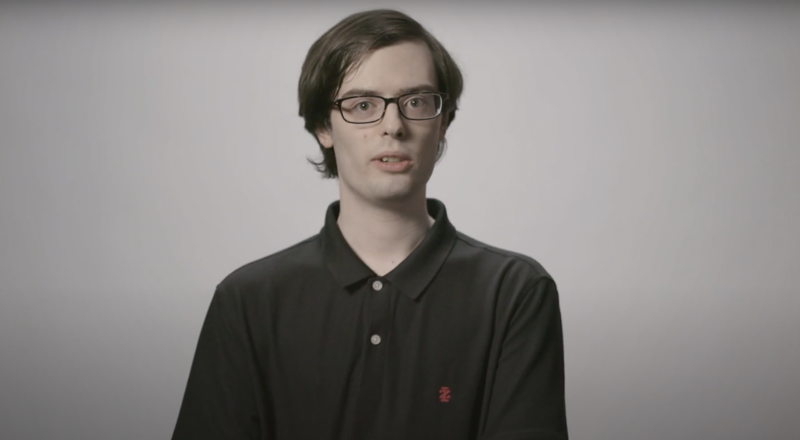 A young man with glasses and straight brown hair, wearing a black polo shirt, stands against a plain light gray background.