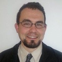 A man with glasses, a goatee, and short dark hair, dressed in a suit and tie, smiles confidently at the camera against a plain background. His poised demeanor reflects his dedication to criminal justice.