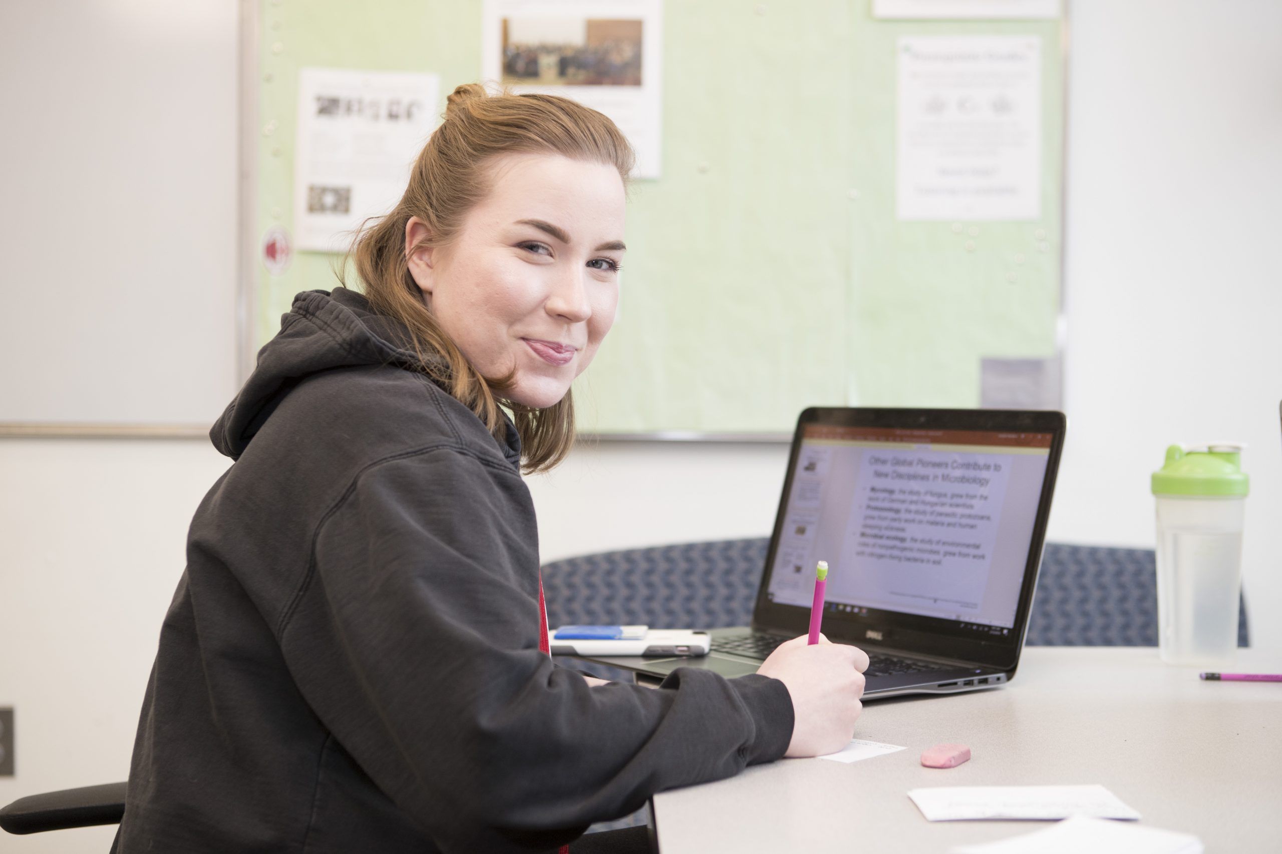 A Worcester state student working on homework on an open laptop, looking into the camera and smiling