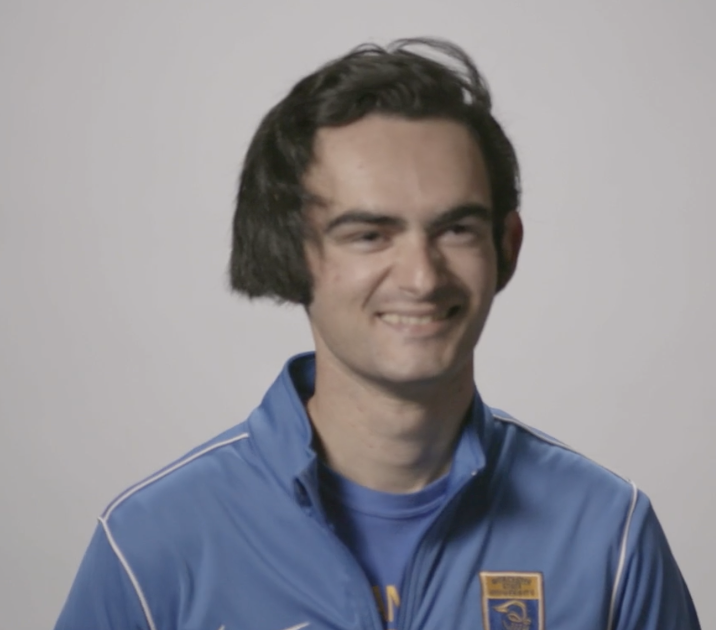 Young man with dark, unevenly cut hair wearing a blue zip-up sports jacket and smiling against a plain light background, embodying the approachable confidence of a Communication Major.