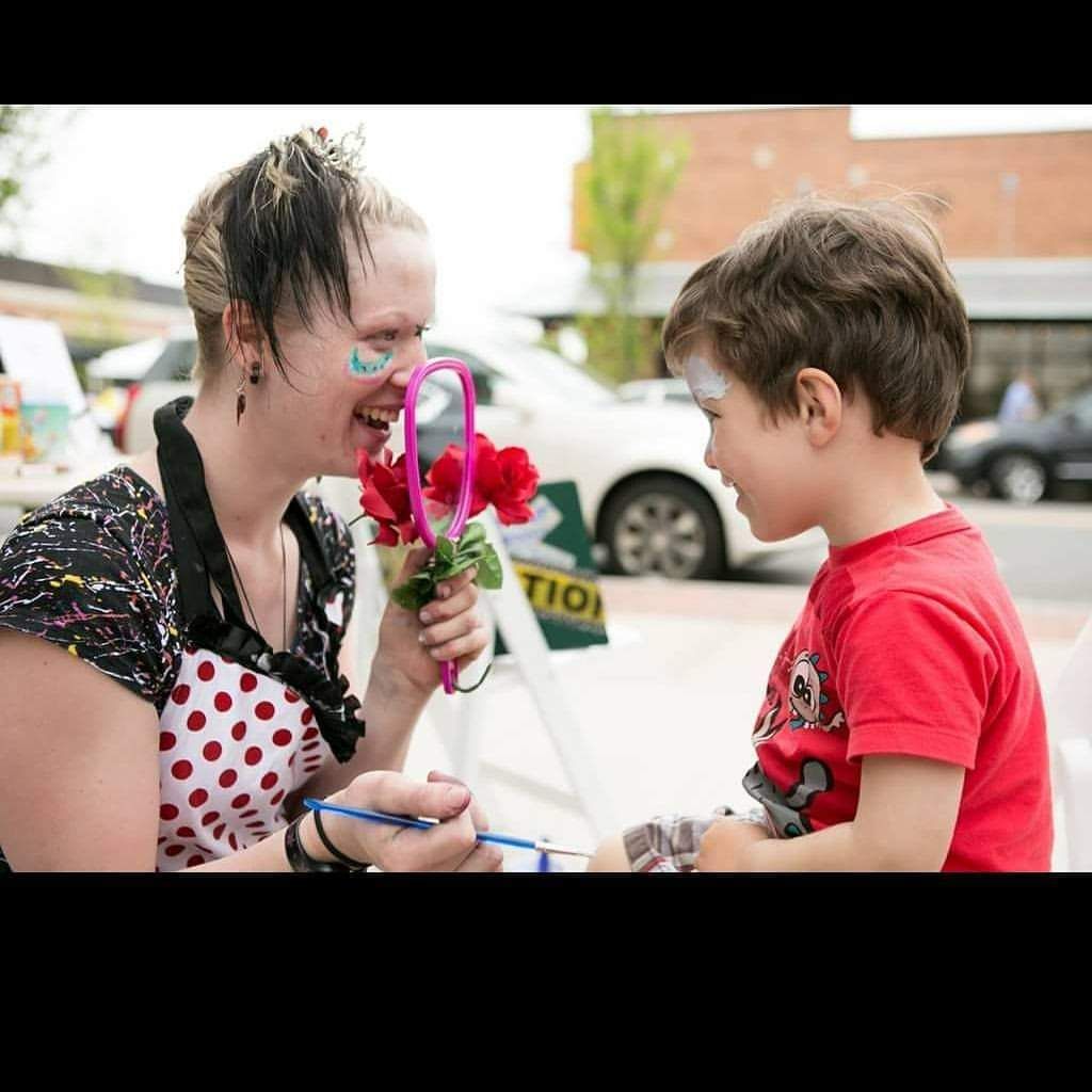 A woman in a polka dot dress, part of the Visual & Performing Arts program, holds a mirror and flowers while face painting a young boy sitting in front of her outdoors.