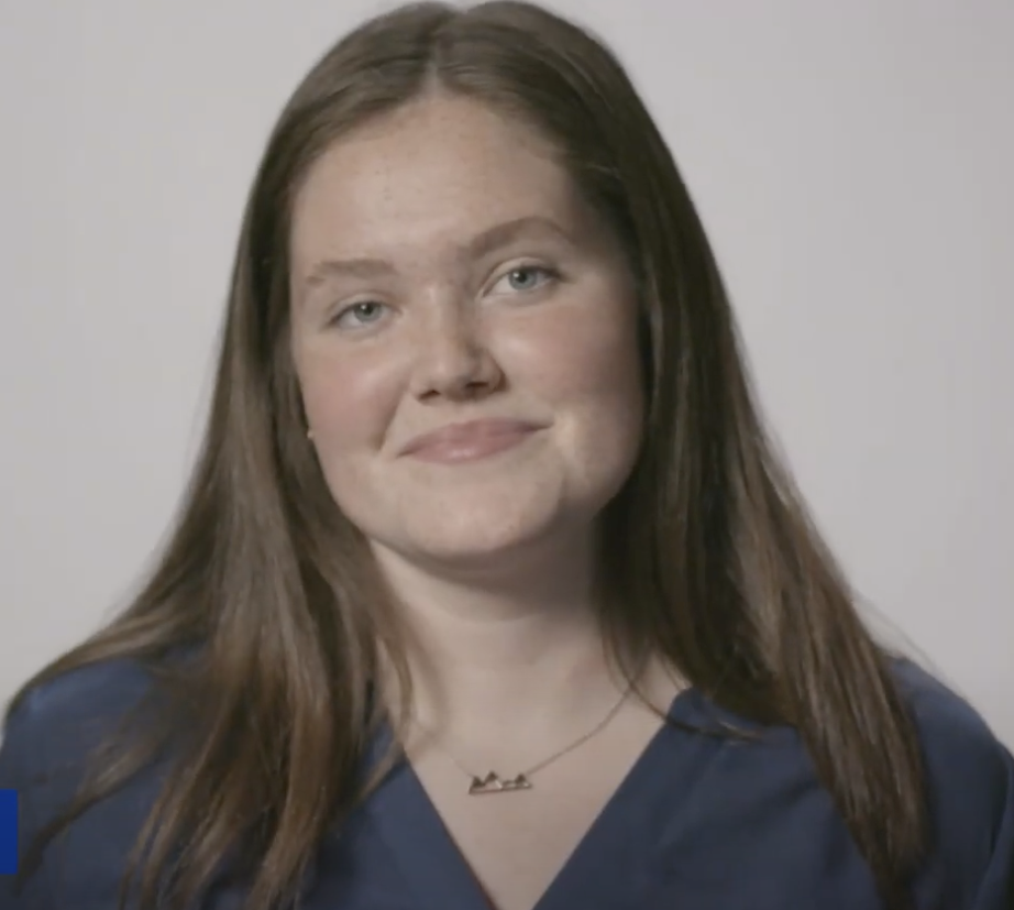 A woman with long brown hair, wearing a blue top and a necklace, smiles slightly as she poses in front of a plain light background, embodying the confidence of a History & Political Science program graduate.