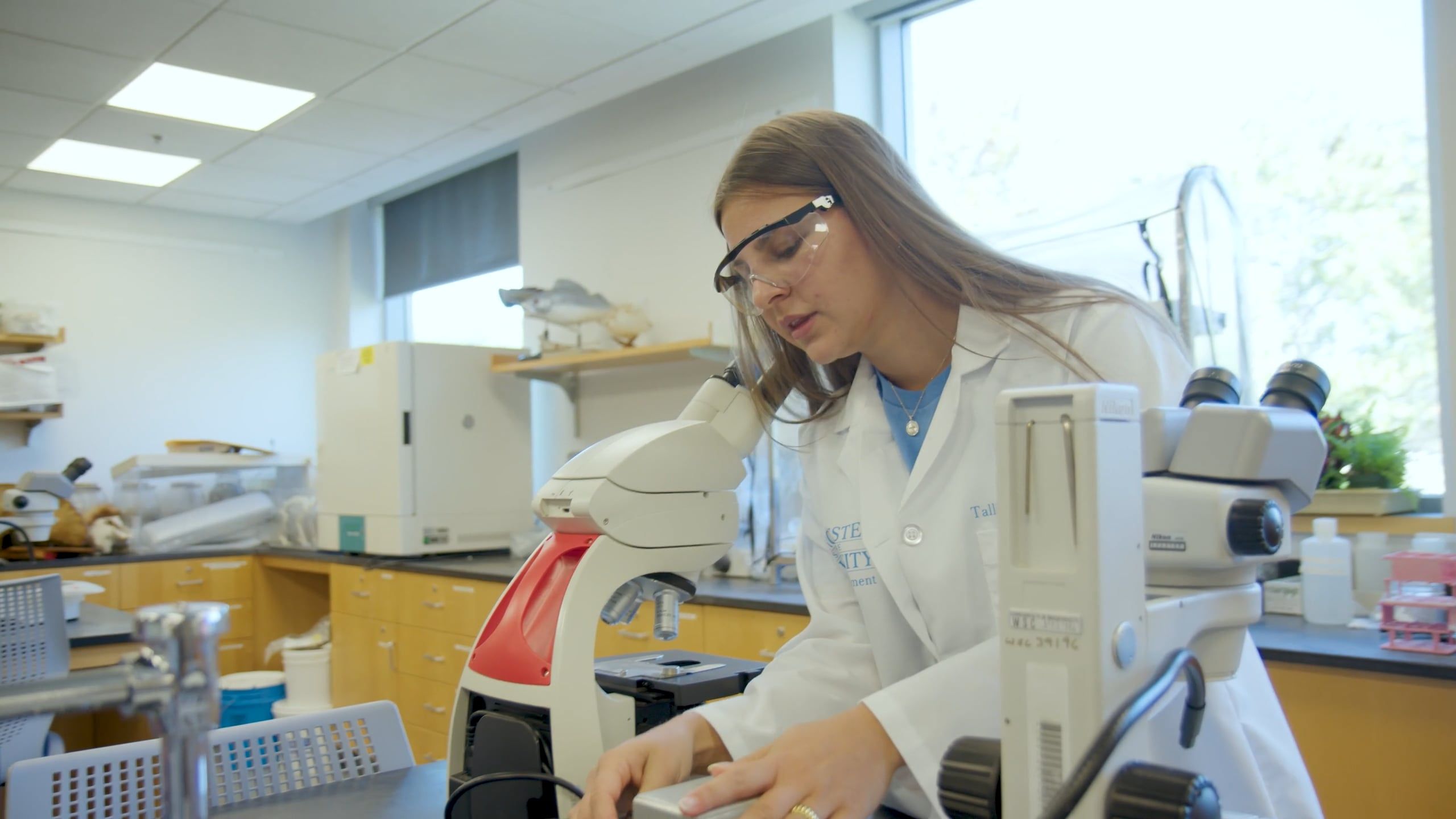 A person in a white lab coat and safety glasses, possibly an Occupational Studies Major, works next to a microscope in a laboratory with scientific equipment on the benches.