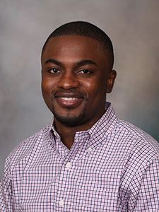 A man with short hair and a trimmed beard is wearing a checkered shirt, smiling at the camera against a neutral background. His confident demeanor perfectly embodies his dedication as a Chemistry major.