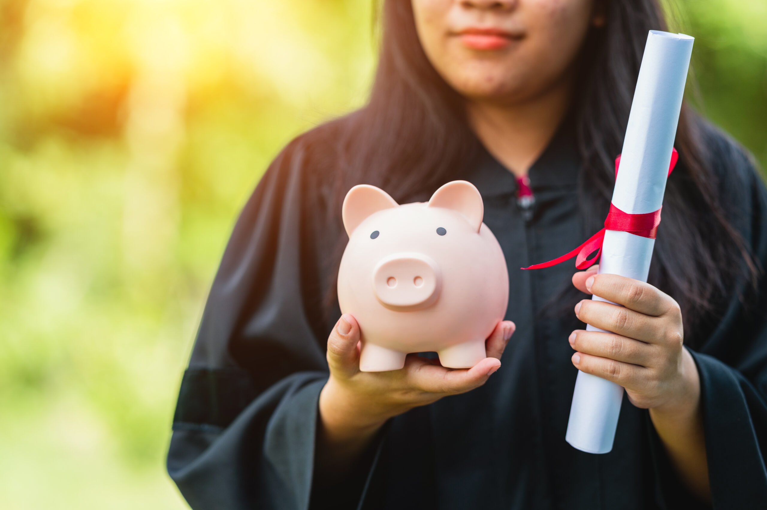 A college graduate holding a diploma in one hand and a piggy bank in the other signifying the costs for a diploma