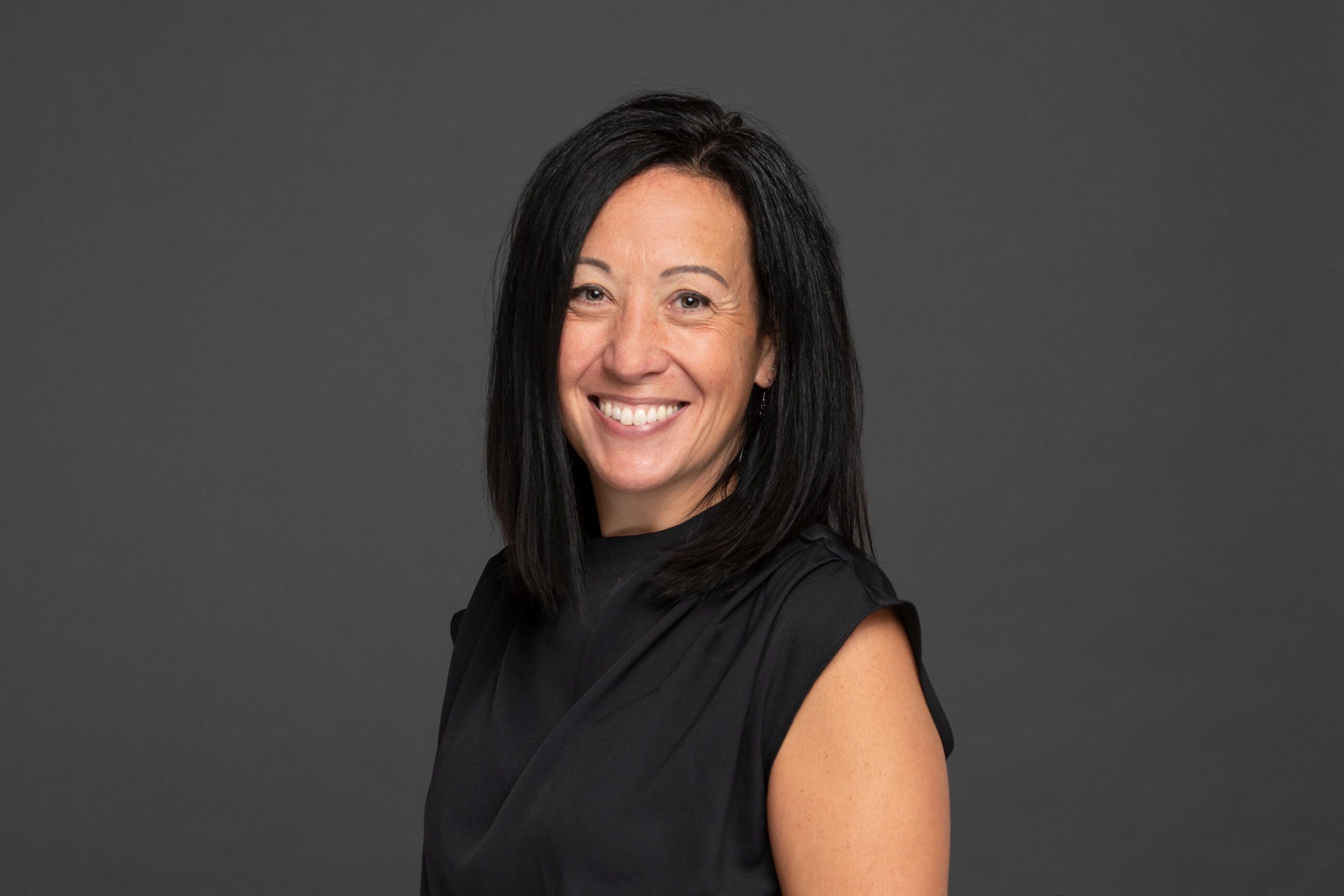 Woman with straight black hair, wearing a sleeveless black top, smiling at the camera against a plain dark gray background.