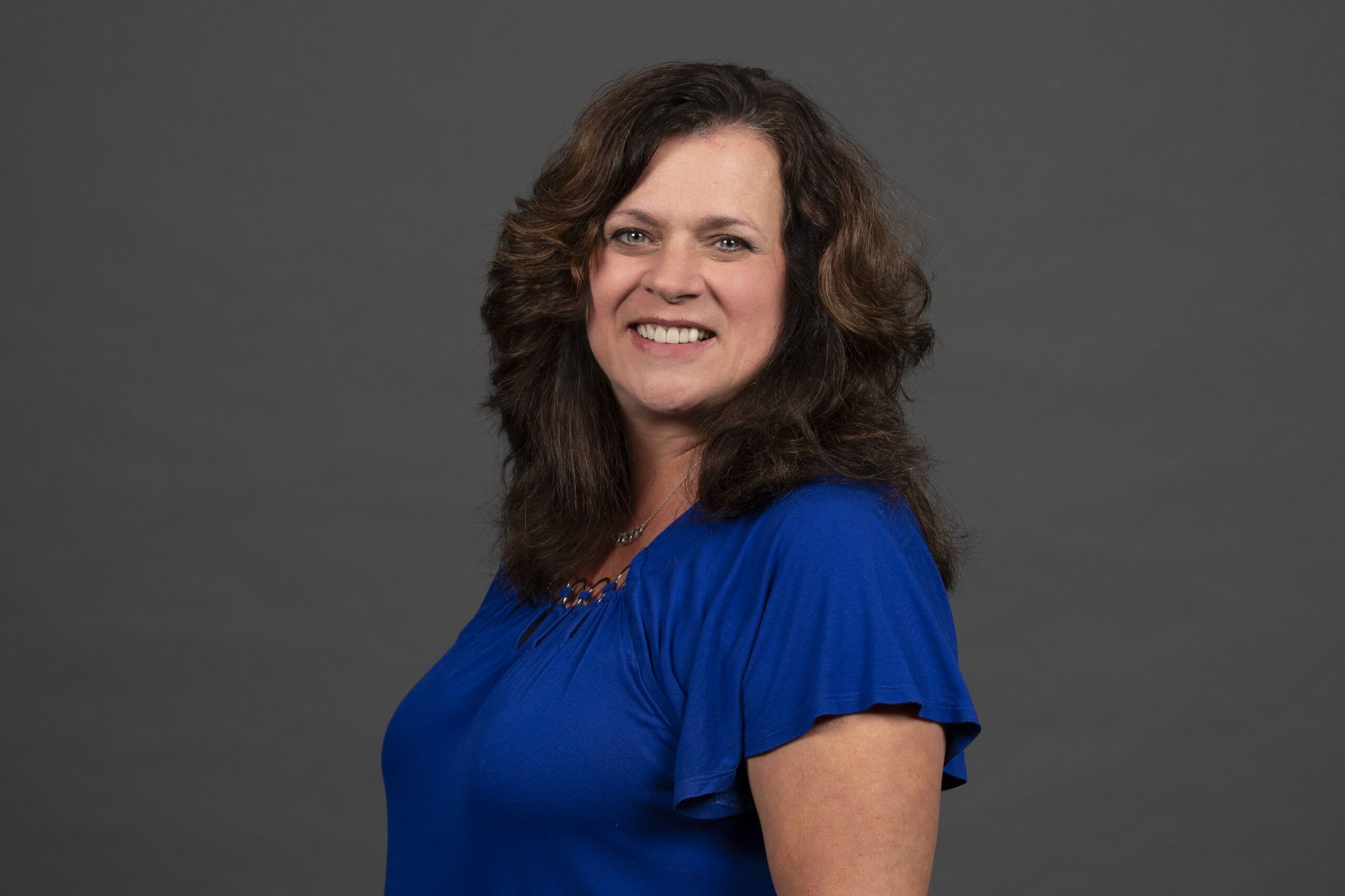 A woman with long curly hair wearing a blue blouse smiles against a gray background.