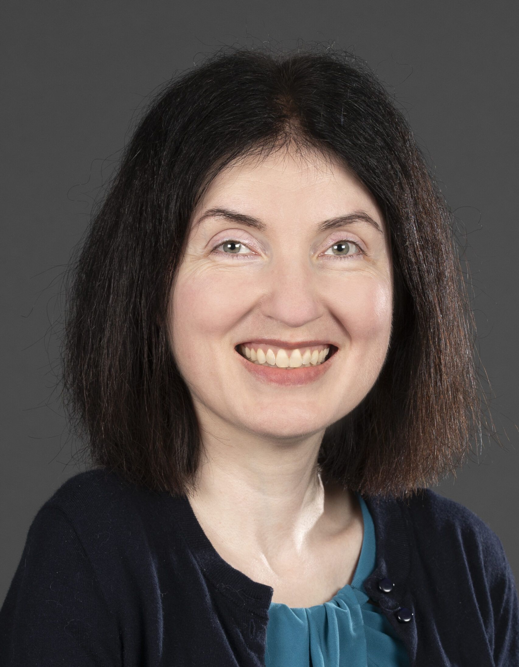 A person with shoulder-length black hair, wearing a teal blouse and dark cardigan, smiles against a gray background.