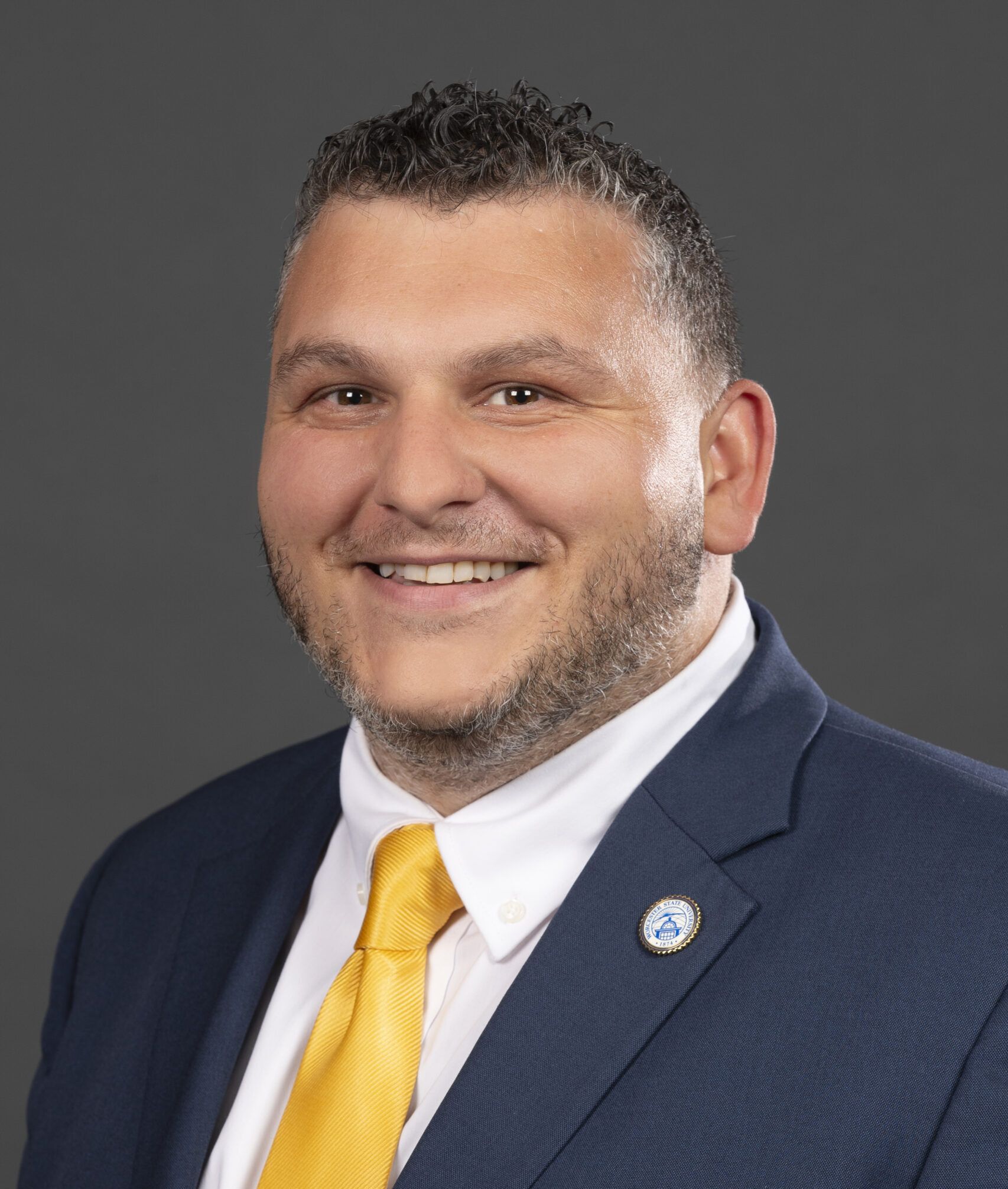 A man in a blue suit and yellow tie smiles against a gray background.