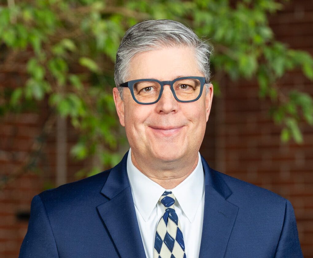 A man in a blue suit and patterned tie stands indoors in front of a brick wall and a leafy green tree, smiling at the camera.