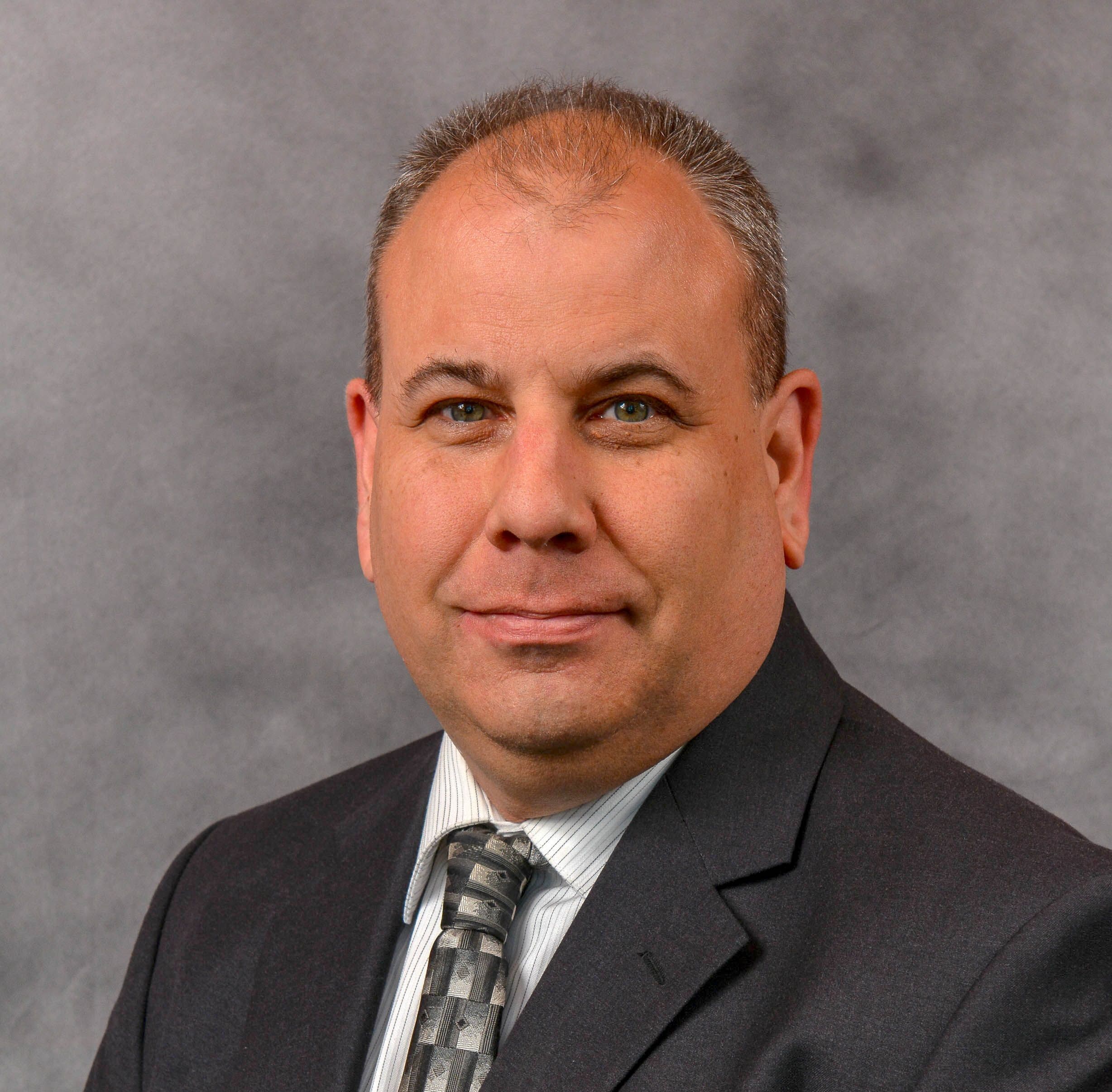 A person in a suit and tie poses against a gray background.