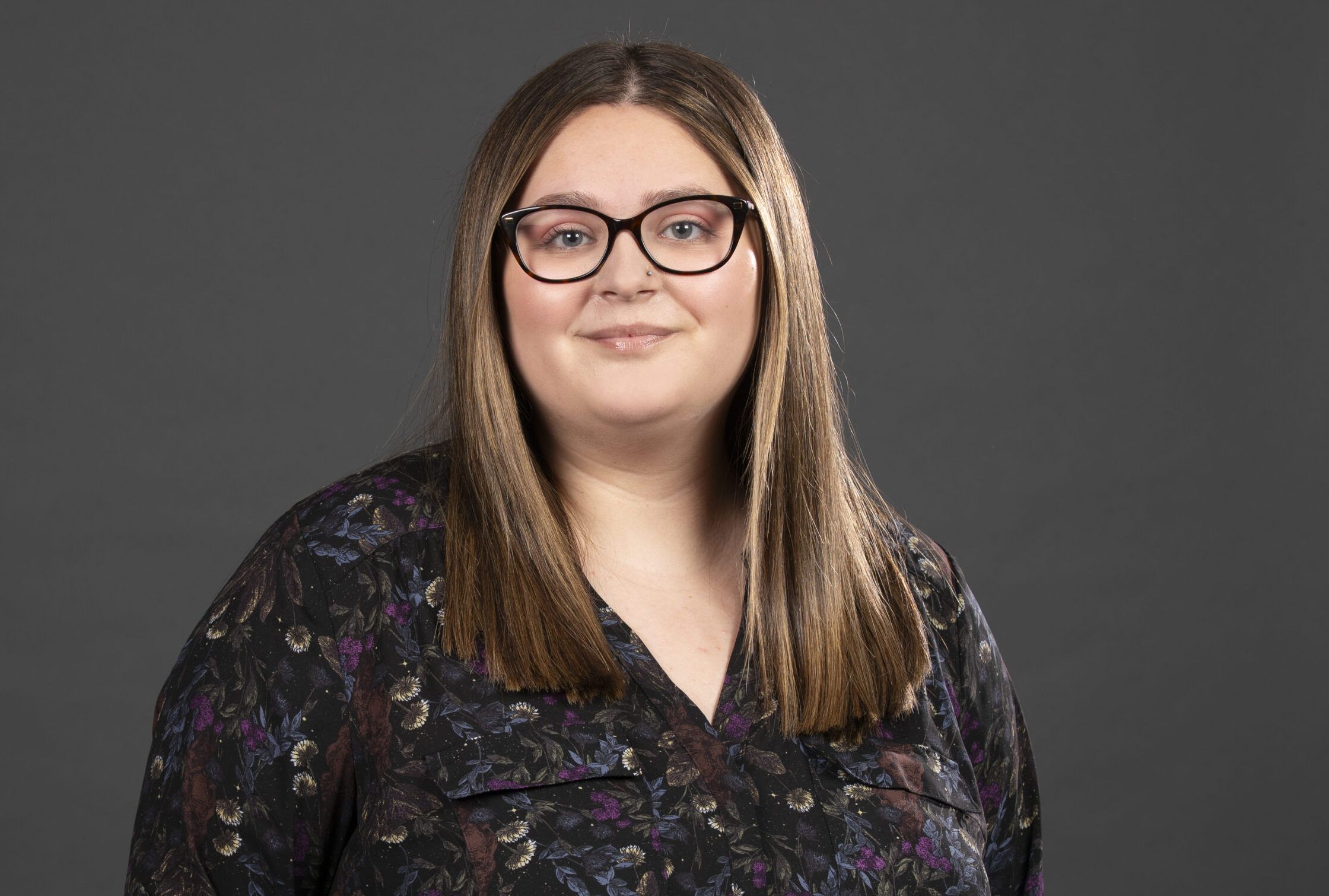 A person with long hair and glasses, wearing a floral-patterned blouse, looking at the camera against a gray background.