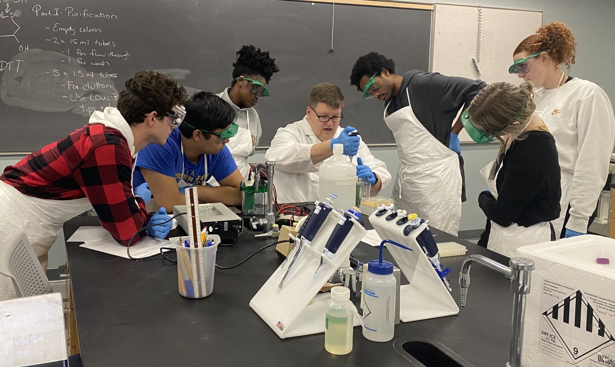 Professor Roger Greenwell sat at a science lab work bench surrounded by students observing his experiment