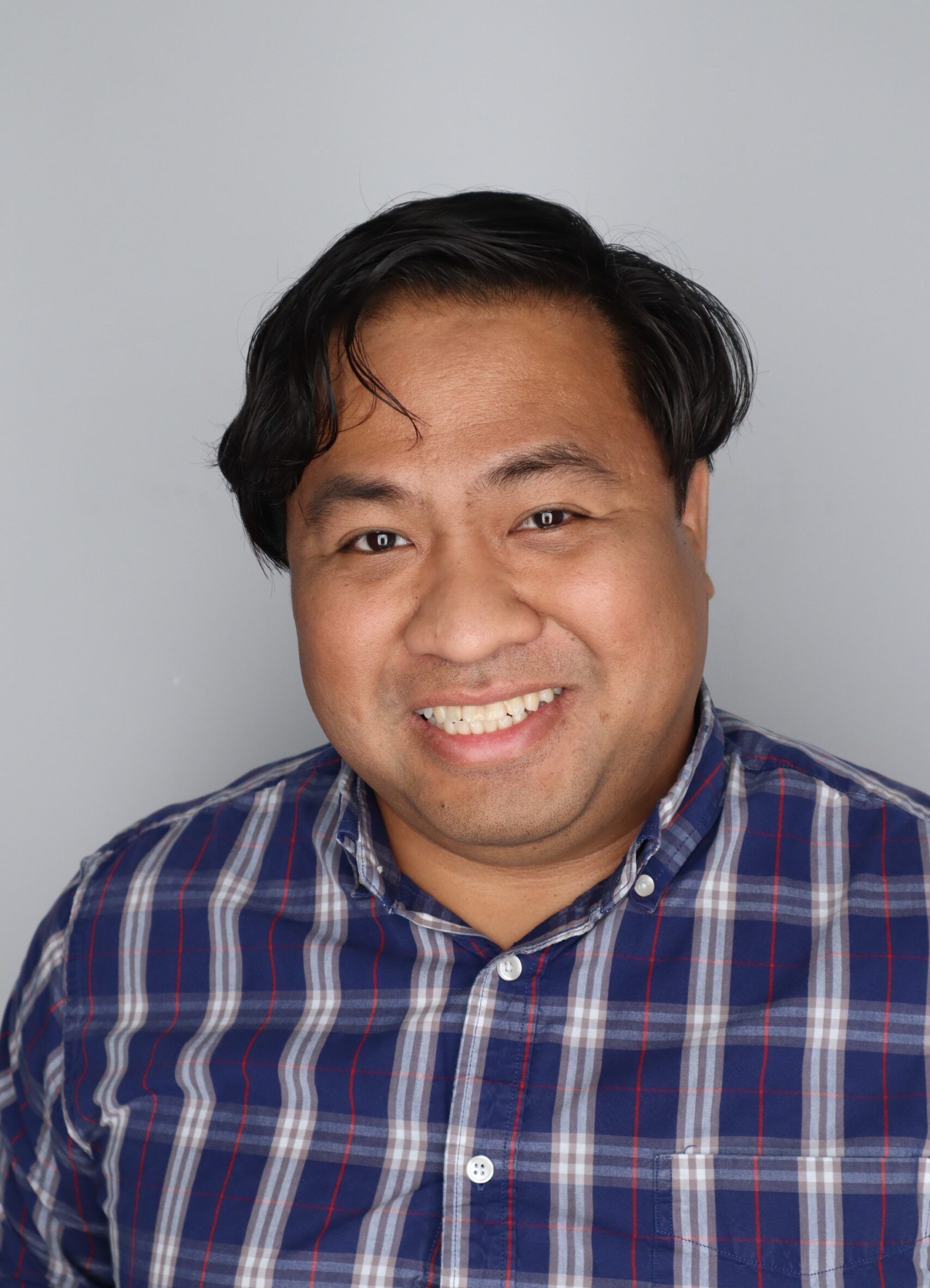 A man with dark hair and a plaid shirt smiles at the camera in front of a plain gray background.
