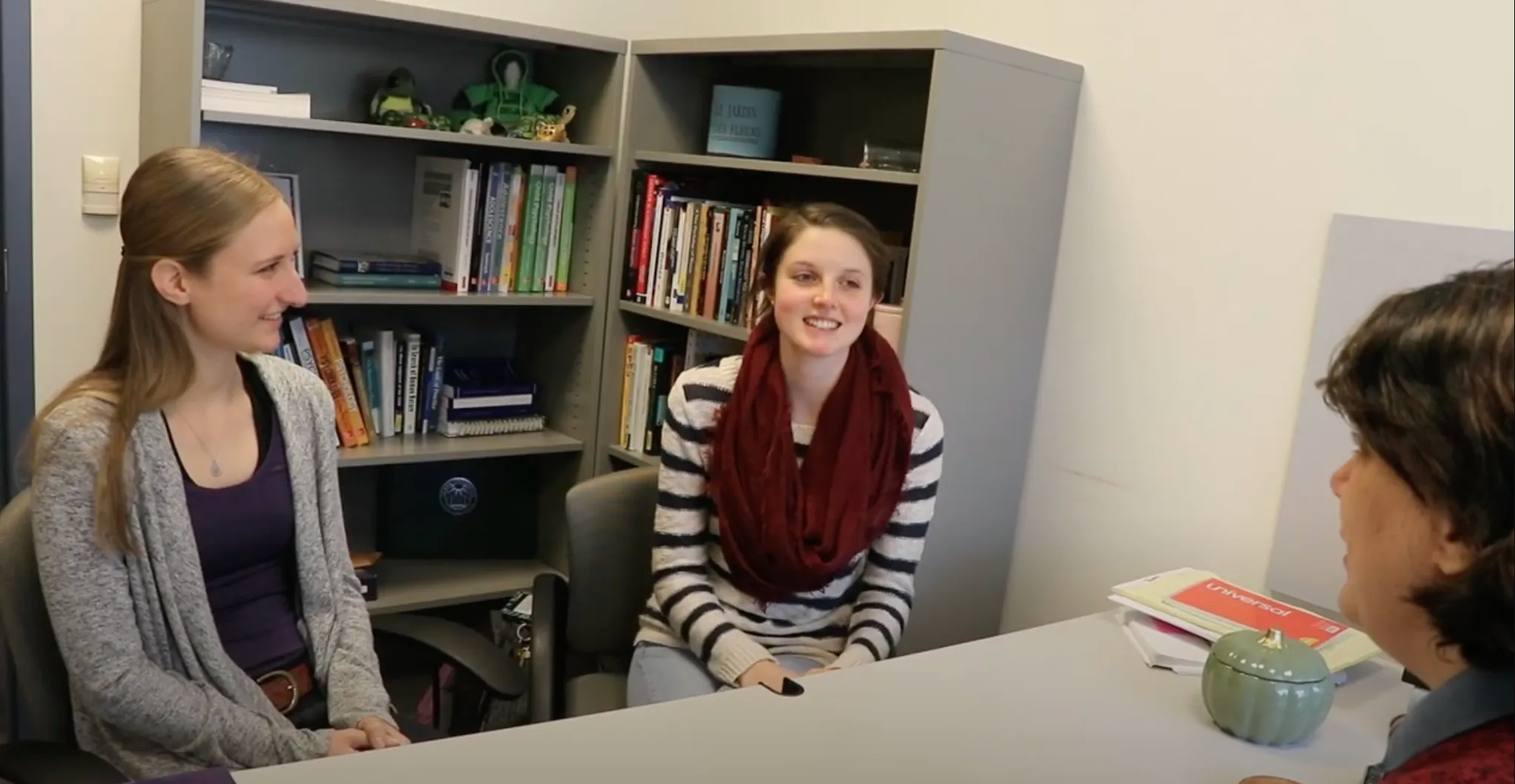 Two women, likely psychology majors, are seated and conversing with another person at a desk in an office. Shelves filled with books and items create a rich backdrop, adding to the academic atmosphere.