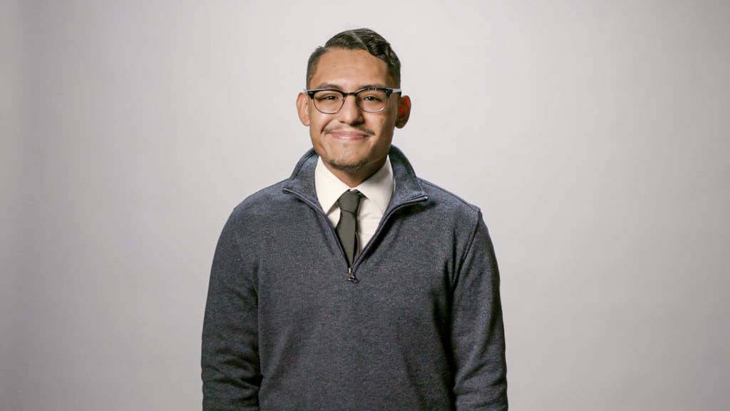 A psychology major with glasses, wearing a navy sweater over a white shirt and black tie, stands against a plain white background, looking directly at the camera and smiling.