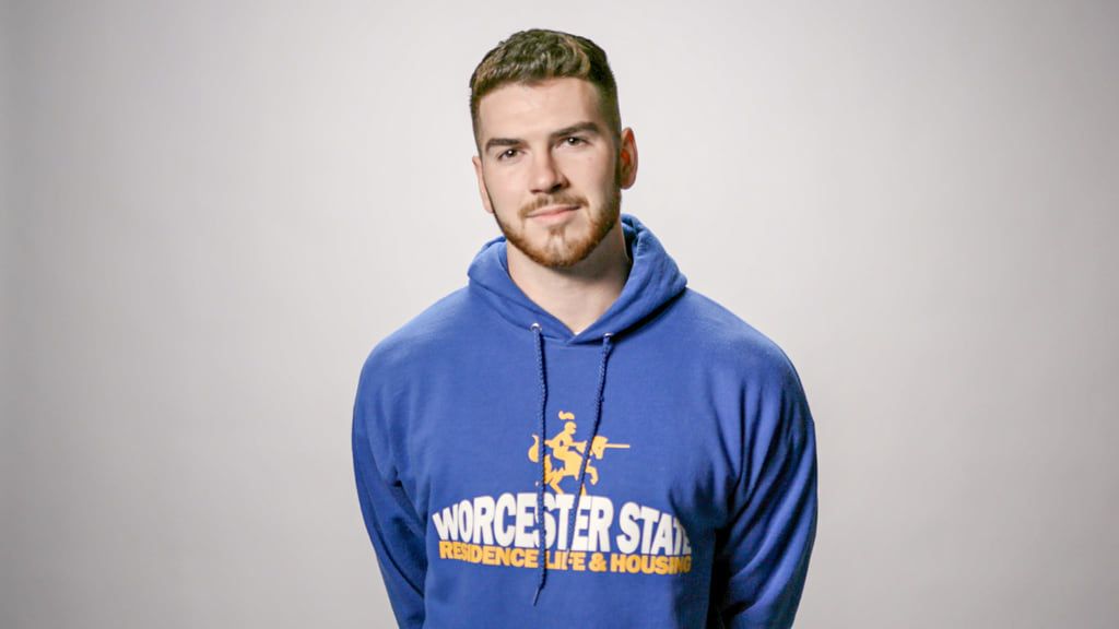A man with short hair and a beard wears a blue hoodie that reads "Worcester State Residence Life & Housing" against a plain background, embodying the laid-back yet thoughtful look of a dedicated Psychology Major.