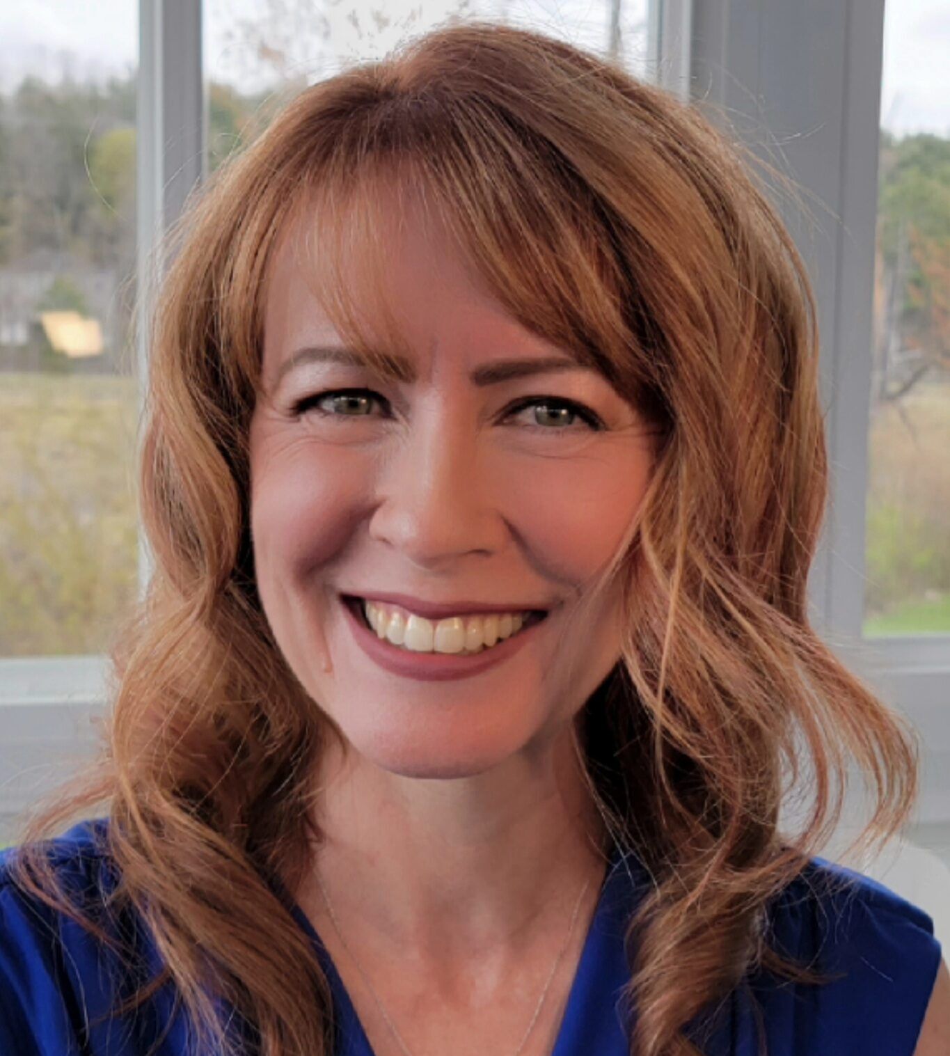 A woman with wavy auburn hair smiles while wearing a blue blouse and a necklace. She is in front of a window with a scenic outdoor background.