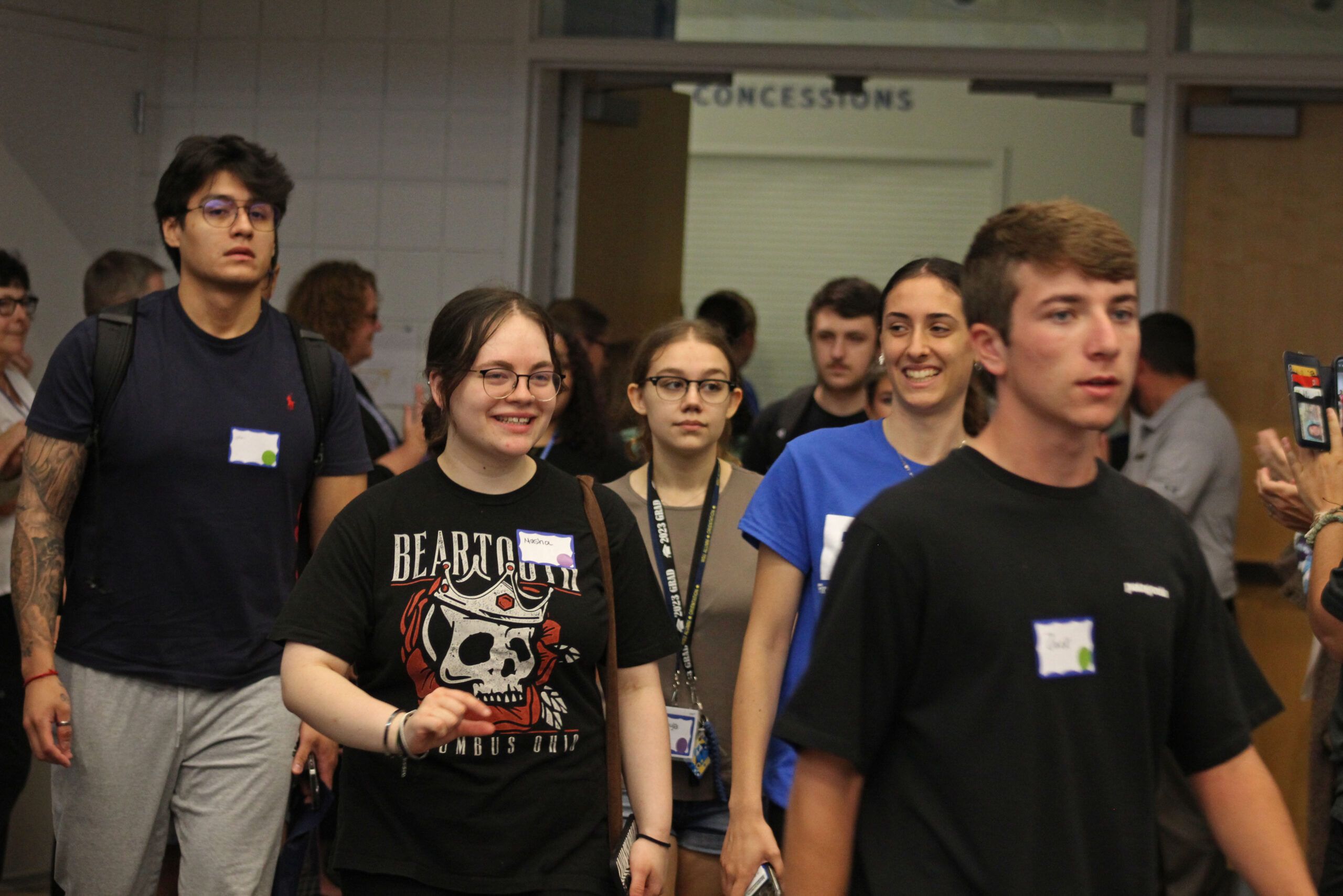 A group of people, mostly young adults, walking indoors. They are wearing casual clothes with name tags visible on their shirts. The sign "Concessions" hangs in the background, suggesting a break during an Academic Convocation event.