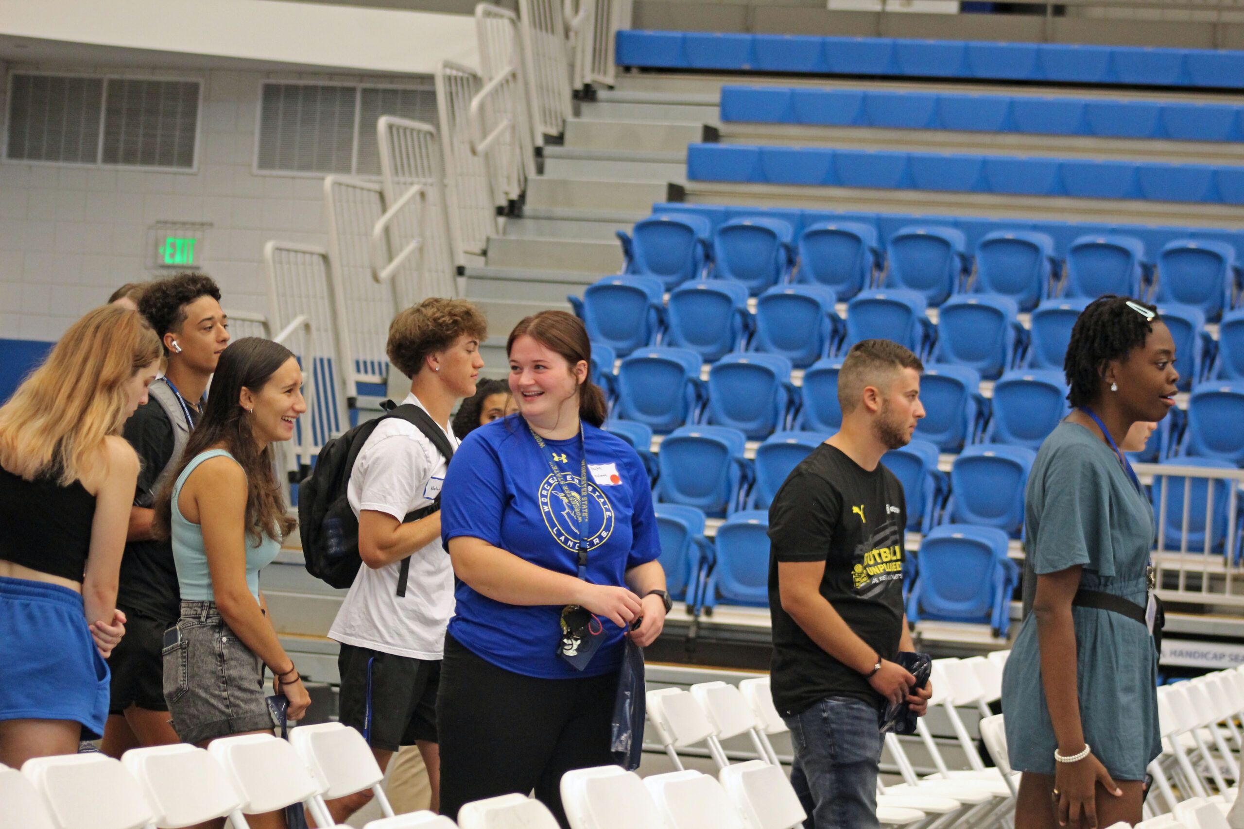 A group of people stands in a gymnasium, some wearing casual clothes and others in sports attire, as they prepare for the Academic Convocation. They are lined up near rows of white folding chairs with blue bleachers in the background.