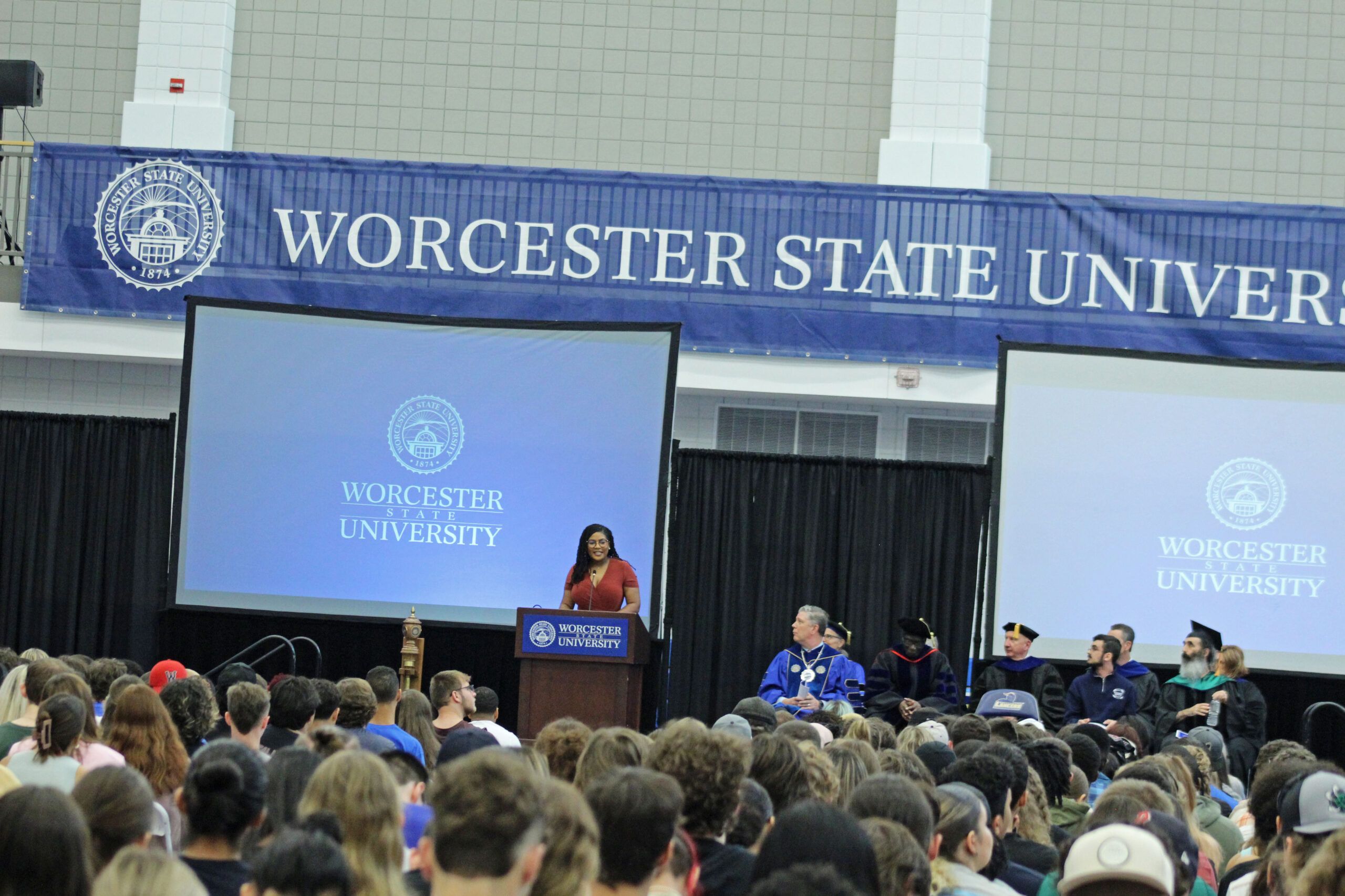 A speaker addresses the audience at Worcester State University's Academic Convocation. Large screens display the university's logo and name, while several seated individuals, some in academic regalia, are on stage.