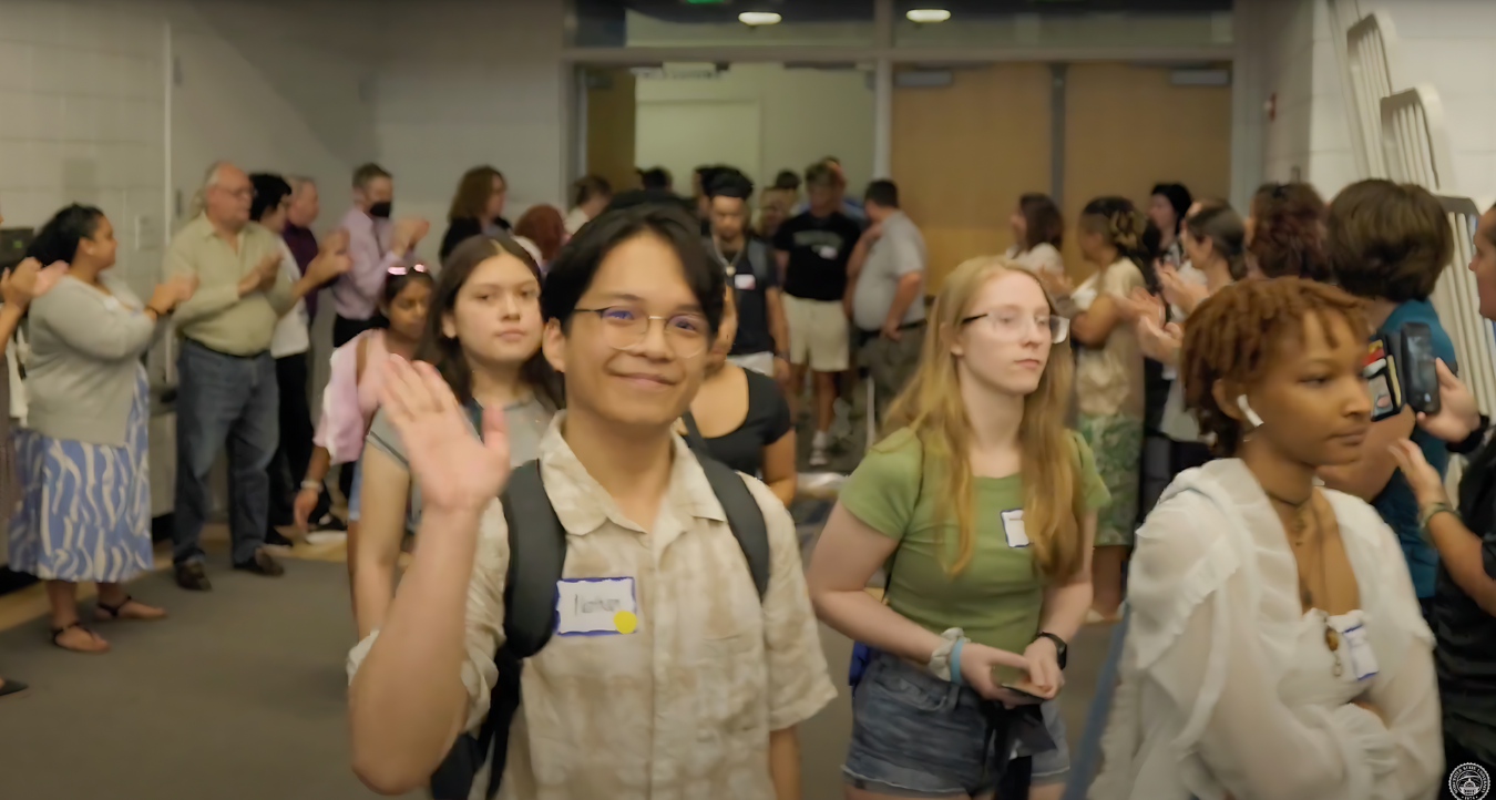 A group of people, mostly young adults with backpacks, walk through a hallway as others stand on either side clapping. One person at the front waves at the camera, capturing the joy of the Academic Convocation moment.