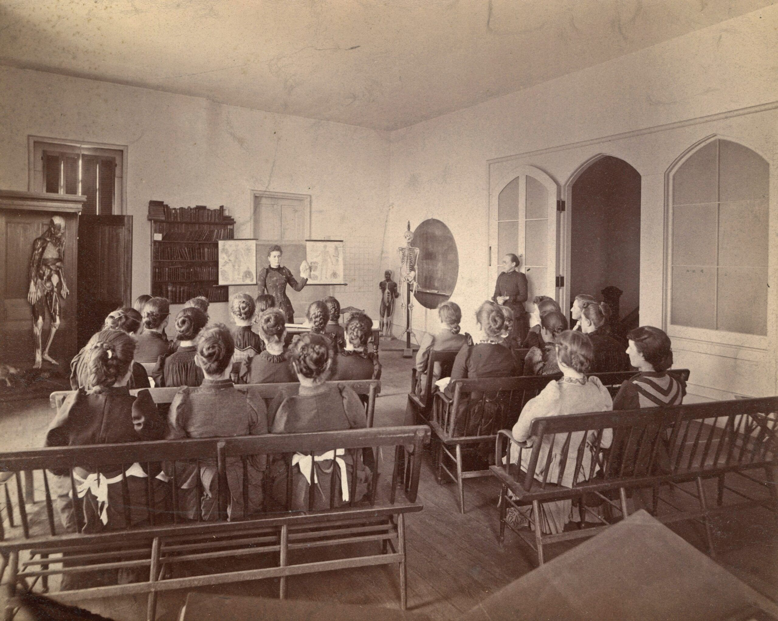 A historical classroom with a group of students seated, attentively listening to an instructor. In celebration of the school's anniversary, various anatomical models and a blackboard with diagrams are prominently displayed at the front of the room.