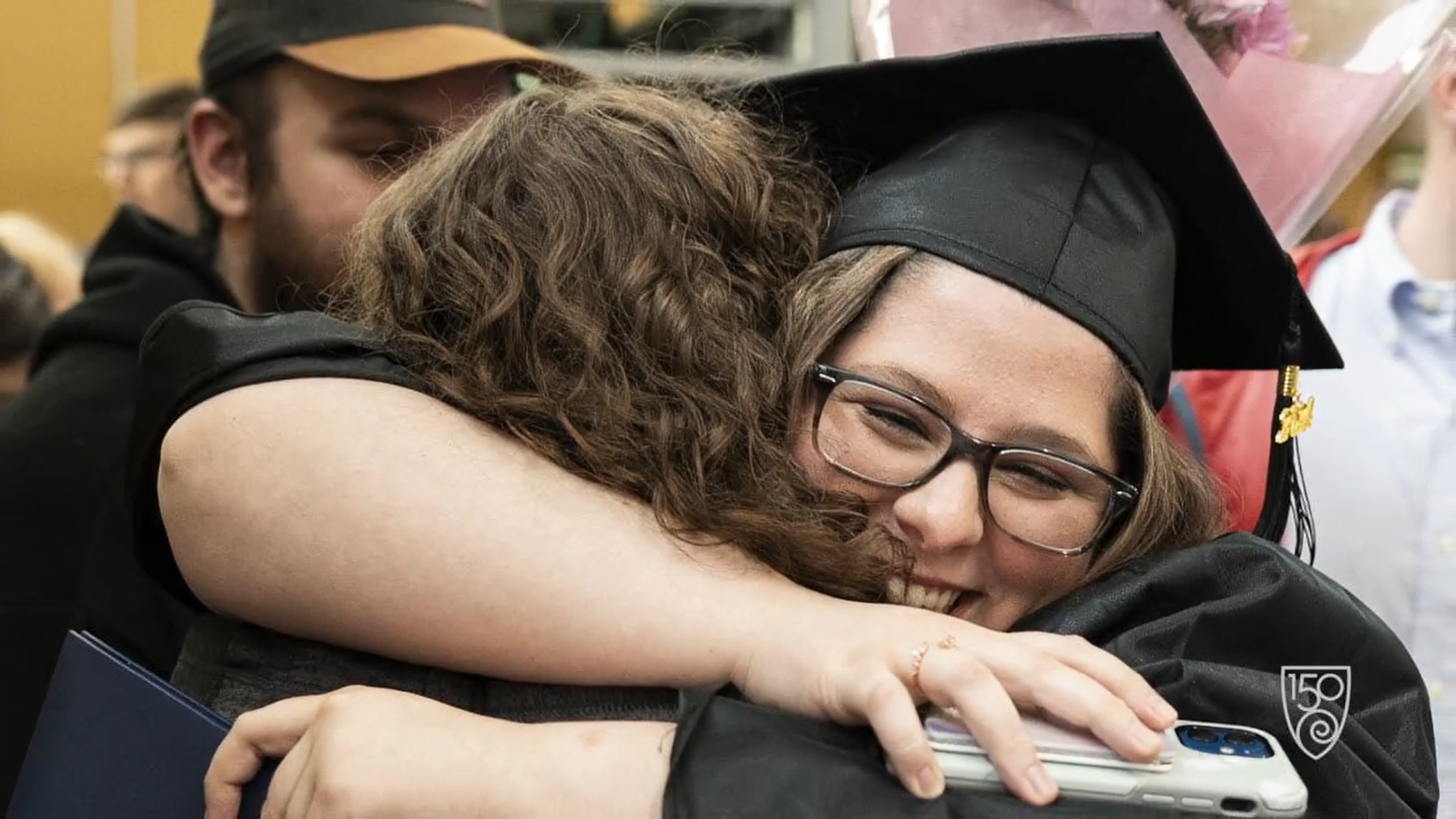 Two people hug at a graduation ceremony, celebrating not just a milestone but an anniversary of hard work and dedication. One wears a cap and gown, holding a phone and diploma, while others look on fondly in the background.