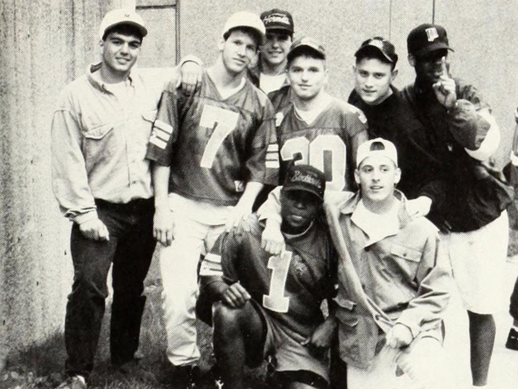 A group of seven people, mostly wearing sports jerseys and caps, pose together outdoors; one person holds up a finger, celebrating an anniversary.